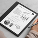 The new Kindle Scribe sitting on a marble countertop while a person holds the Basic pen stylus in their right hand.